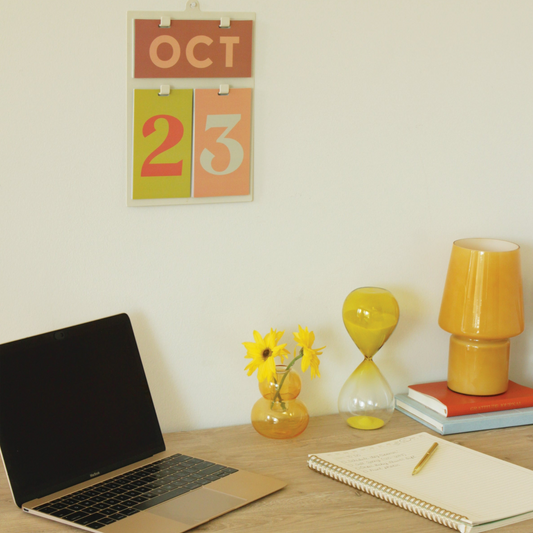 Desk setup with open laptop, notebook, flowers in vase, lamp and a wall calendar mounted above the desk.